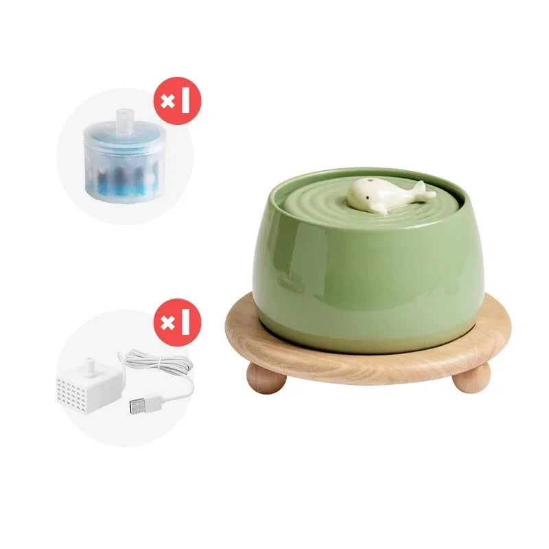 Green ceramic cat water fountain with a motif of a baby whale placed on a wooden stand displayed along with USB charger and replacement filter.