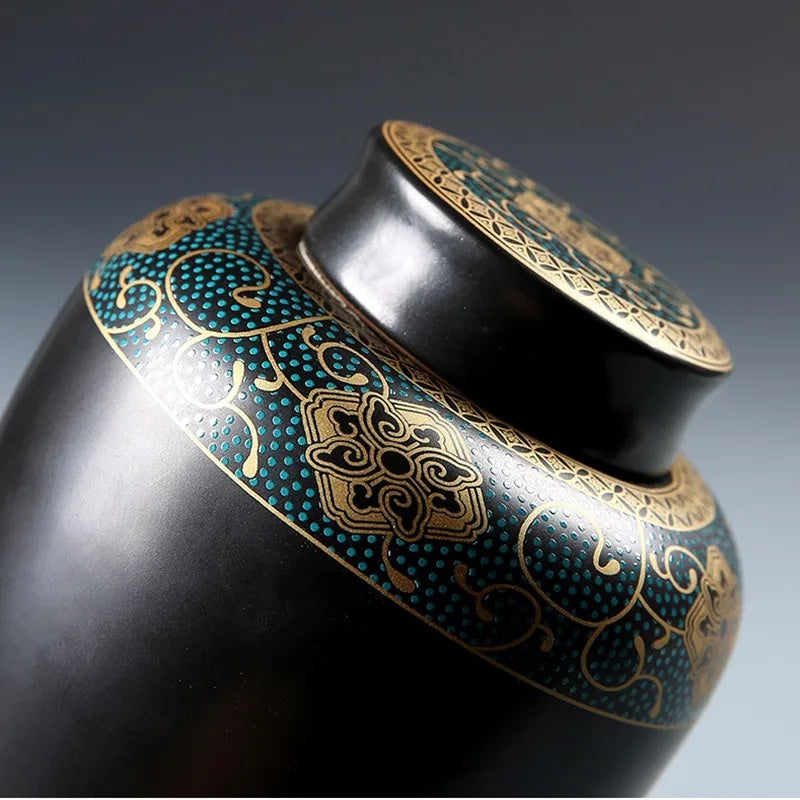 Close up of a ceramic urn for pet ashes with teal and gold ornament on a matte black surface, shown from an angled top view.