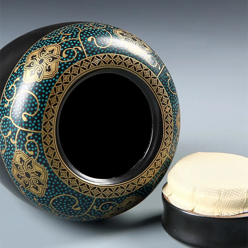 Elegant ceramic urn for pet ashes with teal and gold ornament, shown tilted with open top and matching lid beside it on a light background.