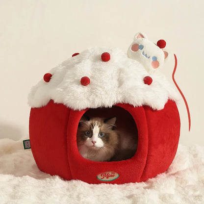 A cat sits inside a red and white cupcake-shaped pet bed with faux-fur top, red cherry details, and a small plush cat on top, set on a white fluffy rug against a light beige background.