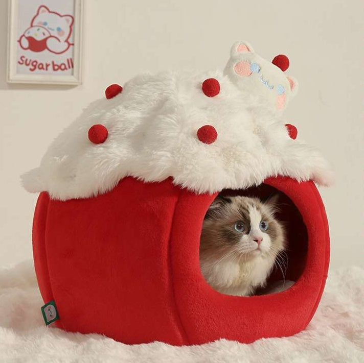 A fluffy white whipped-cream-style roof with red pom-pom details sits on top of a red cat bed. A real cat is visible inside the bed&