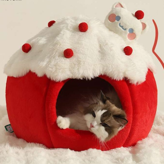 A fluffy cat bed shaped like a red pumpkin or pot with a white faux-fur roof and red pom-pom accents, with a cat resting inside. A small plush cat face is attached near the top on a clean light background.