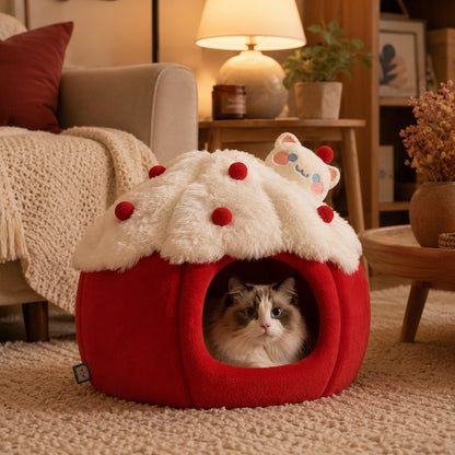A fluffy red and cream cat bed shaped like a cupcake sits on a beige carpet in a cozy living room. A white cat is nestled inside the bed, and the background features a warm lamp, sofa, and wooden furniture.