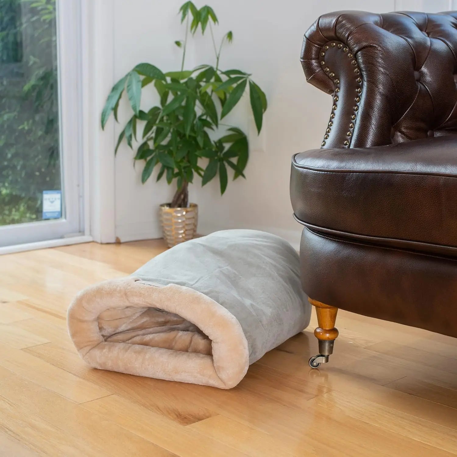 Cat sleeping bag on a wooden floor next to a brown leather sofa with a plant in the background.